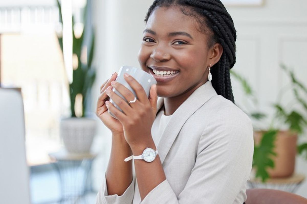 A smiling young woman with braided hair, wearing a light-colored blazer and watch, holding a grey coffee mug with both hands. She is sitting in a modern, well-lit office space with green plants in the background.