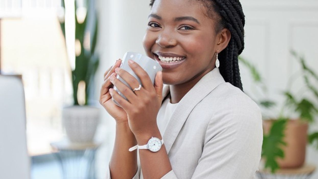 A smiling young woman with braided hair, wearing a light-colored blazer and watch, holding a grey coffee mug with both hands. She is sitting in a modern, well-lit office space with green plants in the background.