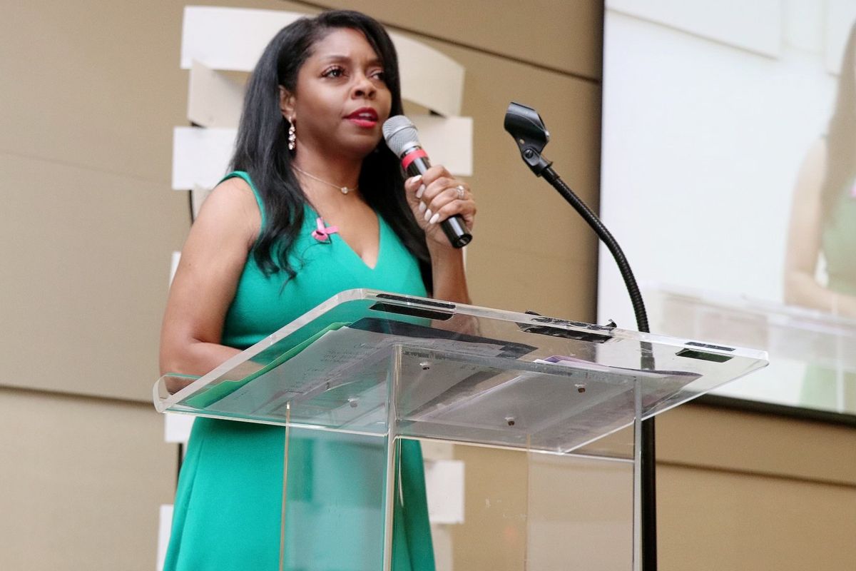 A woman in a sleeveless green dress stands at a clear podium, speaking into a handheld microphone. She has long, dark hair and wears a pink ribbon pin, symbolizing breast cancer awareness. A projection screen behind her shows a live feed of her speech.