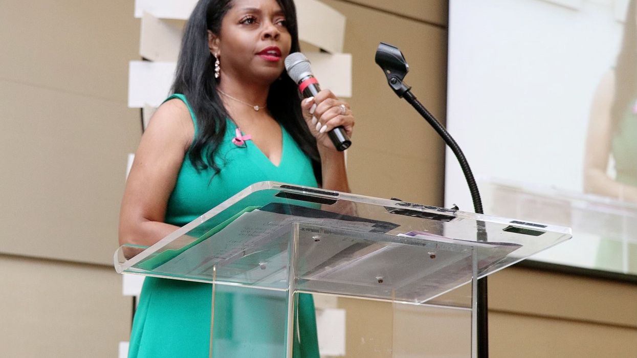 A woman in a sleeveless green dress stands at a clear podium, speaking into a handheld microphone. She has long, dark hair and wears a pink ribbon pin, symbolizing breast cancer awareness. A projection screen behind her shows a live feed of her speech.
